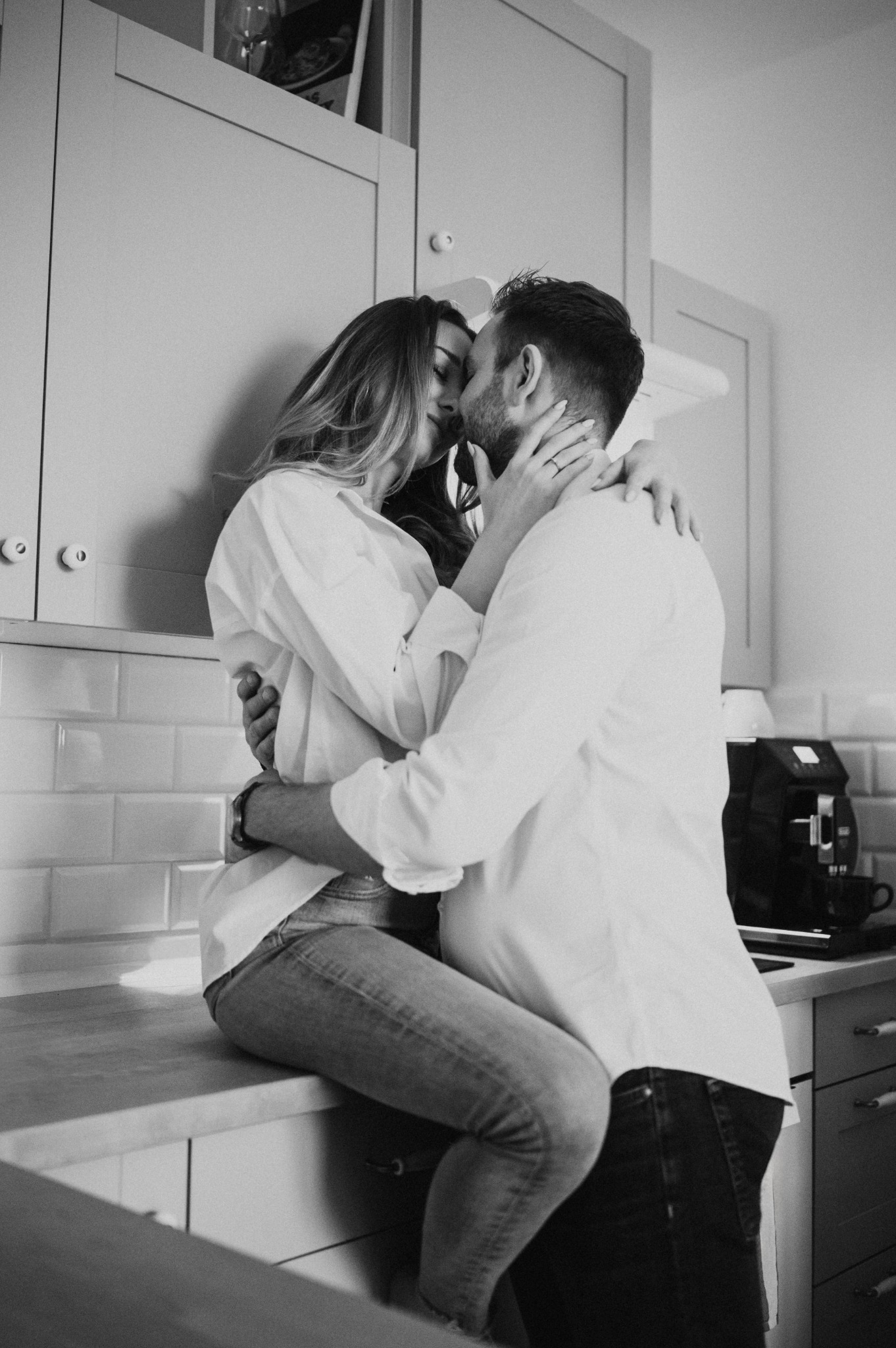 Czarno-białe zdjęcie całującej się pary w kuchni, kobieta siedzi na blacie, oboje ubrani w białe koszule.