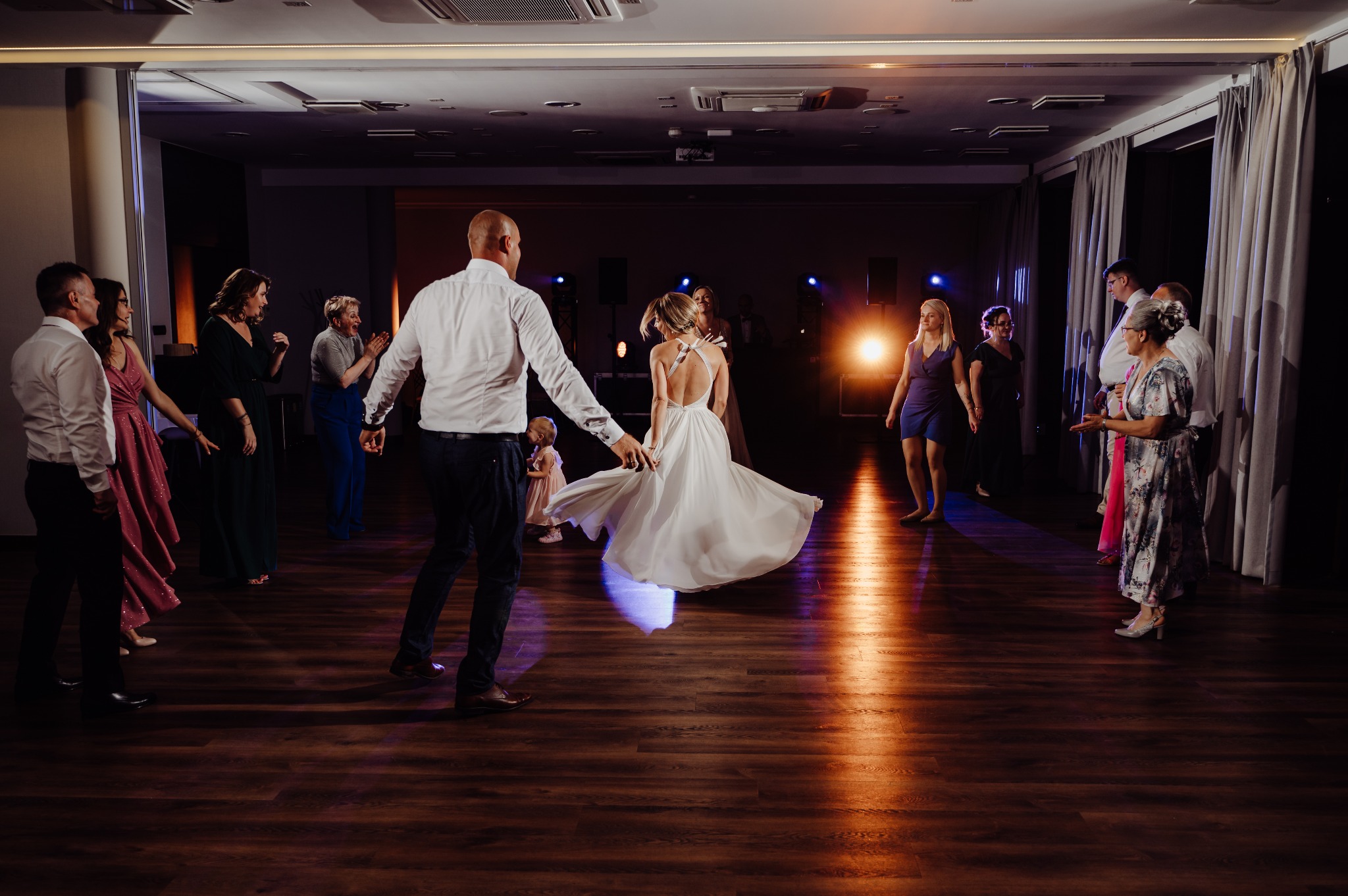 Para młoda tańczy na parkiecie podczas wesela, goście obserwują, widoczne oświetlenie sceniczne.