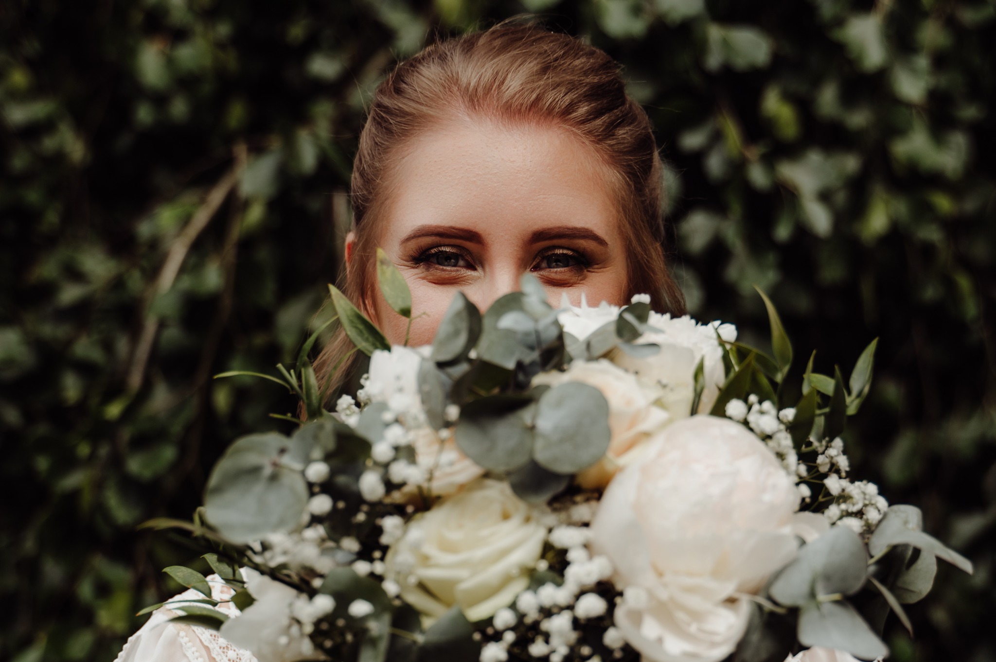 Uśmiechnięta panna młoda, z widocznymi oczami znad białego bukietu ślubnego na tle zieleni. Delikatny makijaż podkreśla spojrzenie.