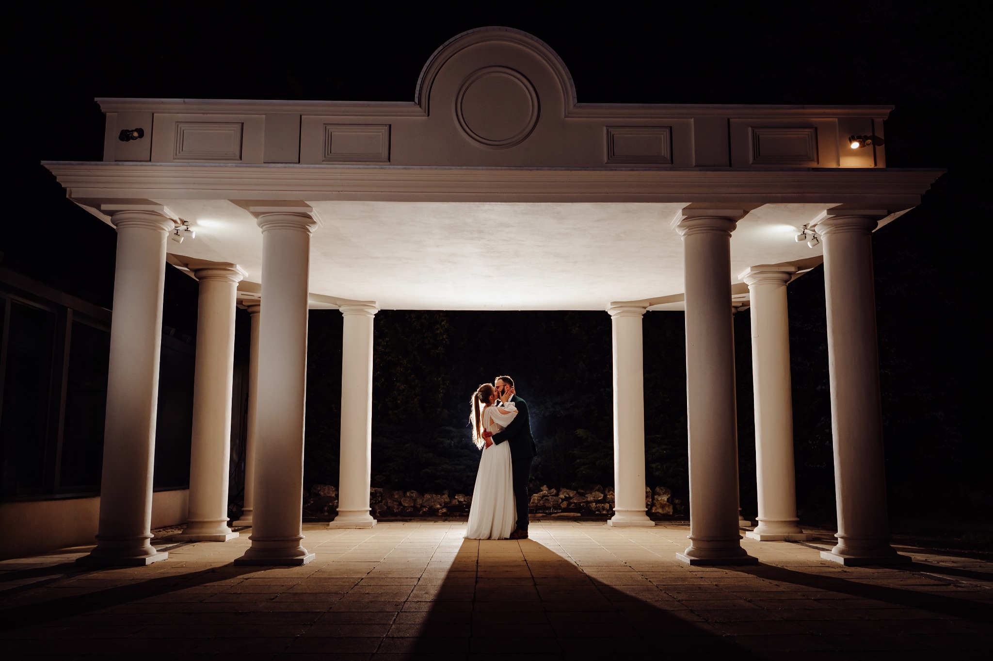 Para młoda w objęciach pod oświetlonym, kolumnowym pawilonem w nocnej scenerii, z widocznym blaskiem światła podkreślającym ich sylwetki.