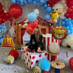 Animator w czarnym stroju siedzi na kostce z czerwonymi kropkami, otoczona dekoracjami cyrkowymi, balonami i konfetti w kształcie gwiazdek.