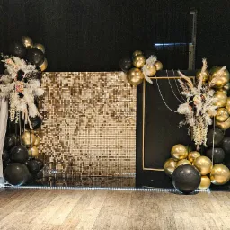 Dekoracja eventowa: błyszcząca ścianka z mozaiki, kompozycje z czarnych i złotych balonów, piór oraz kwiatów, podświetlenie LED na podłodze.