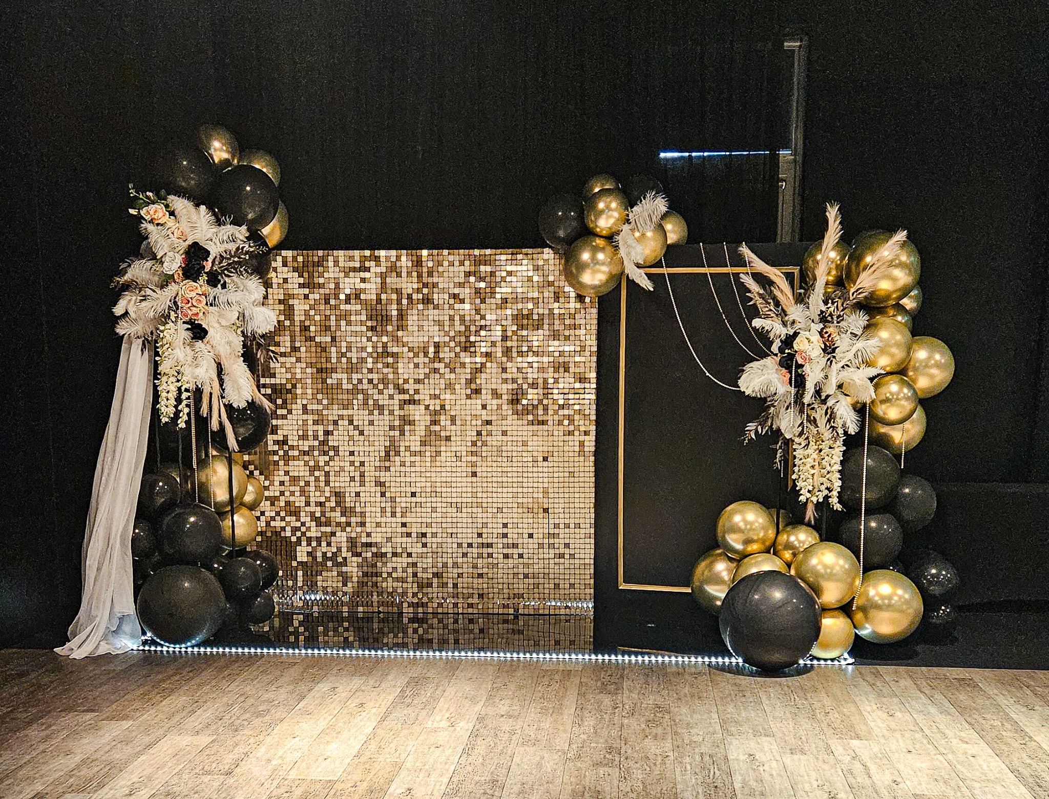 Dekoracja eventowa: błyszcząca ścianka z mozaiki, kompozycje z czarnych i złotych balonów, piór oraz kwiatów, podświetlenie LED na podłodze.