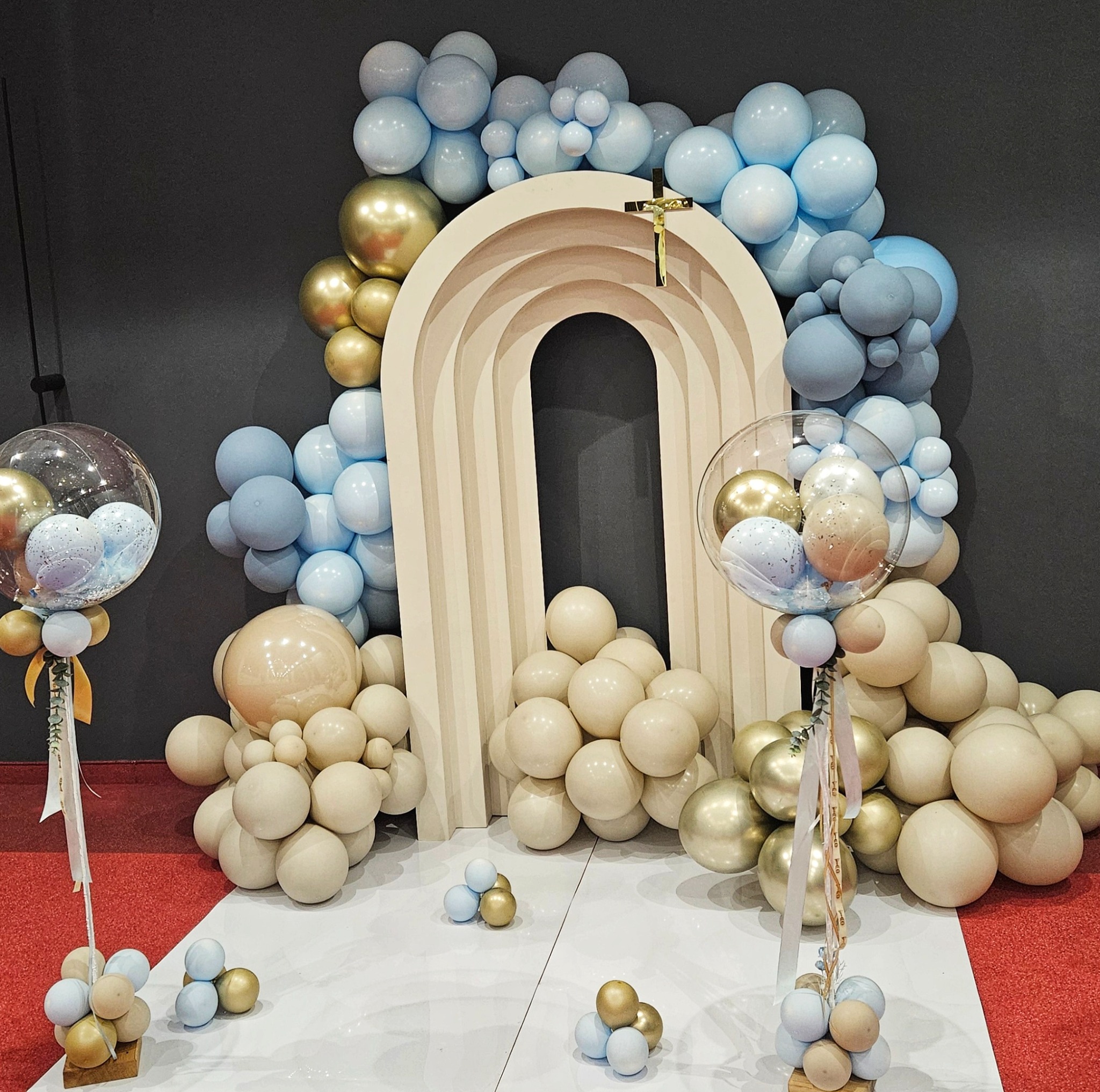 Dekoracja z balonów w odcieniach błękitu, złota i beżu, z drewnianą konstrukcją w kształcie łuku i złotym krzyżem, na białej podłodze z czerwonym dywanem w tle.