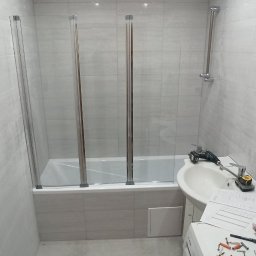 BudDom - Łazienka w trakcie montażu szklanego parawanu nawannowego, widoczne narzędzia, umywalka, pralka i płytki ścienne.