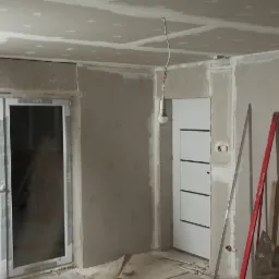 Wnętrze w trakcie prac wykończeniowych: ściany pokryte płytami gipsowo-kartonowymi, widoczne łączenia szpachlowane, sufit z zamontowanymi płytami GK, wisząca żarówka na kablu, drzwi wewnętrzne...