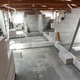 Poddasze w trakcie adaptacji, z widocznymi ścianami z białych bloczków, drewnianą konstrukcją dachu i surową, betonową podłogą. Na podłodze widoczne resztki materiałów budowlanych i stos bloczków.