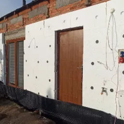 Budynek w trakcie ocieplania styropianem, widoczne okno, drzwi, gniazdka elektryczne i czarna folia fundamentowa.