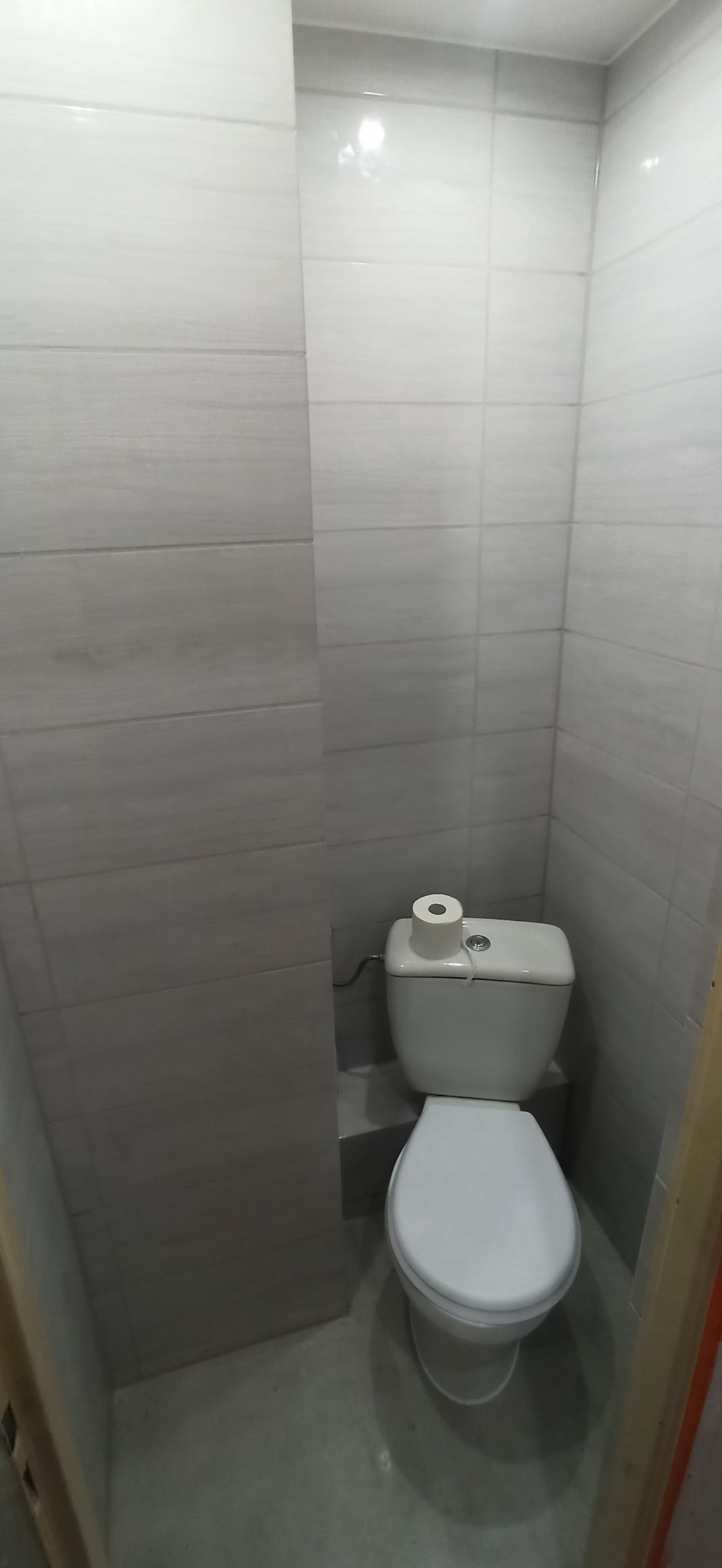 Mała łazienka z toaletą, wykończona szarymi, poziomymi płytkami imitującymi drewno. Na spłuczce rolka papieru toaletowego.