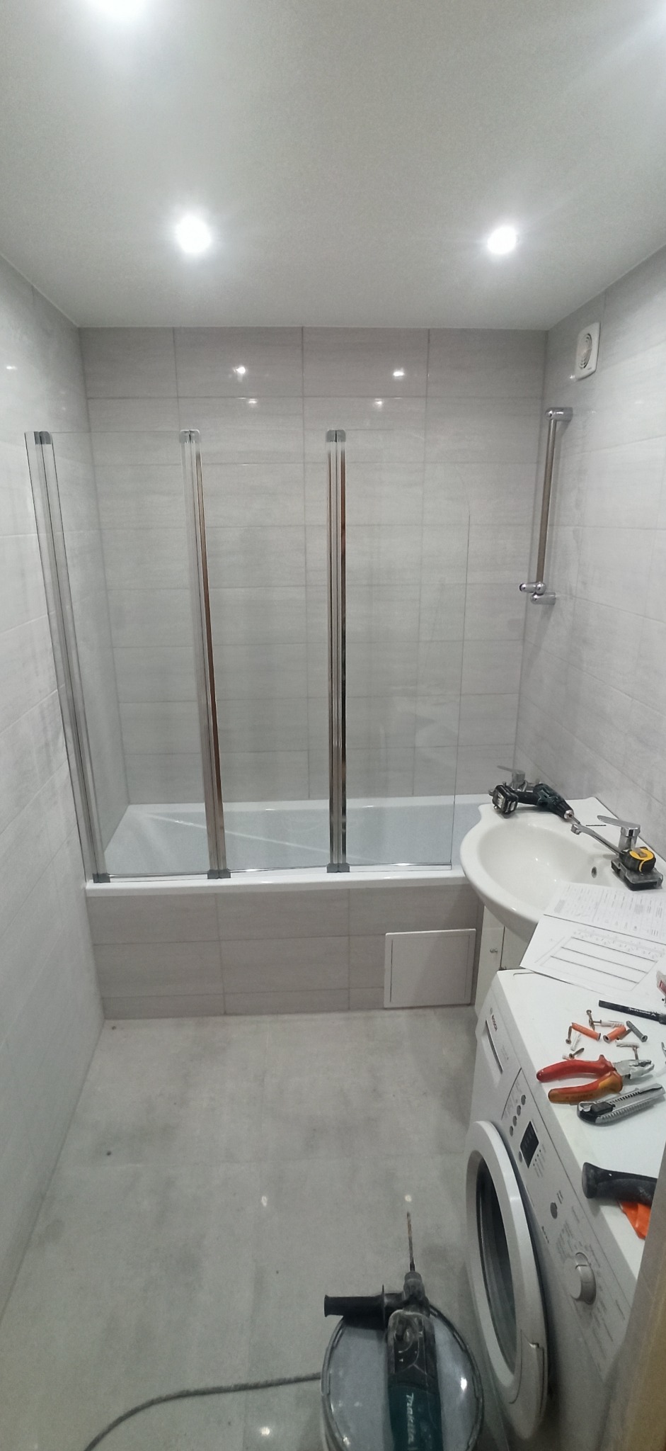 Łazienka w trakcie montażu szklanego parawanu nawannowego, widoczne narzędzia, umywalka, pralka i płytki ścienne.