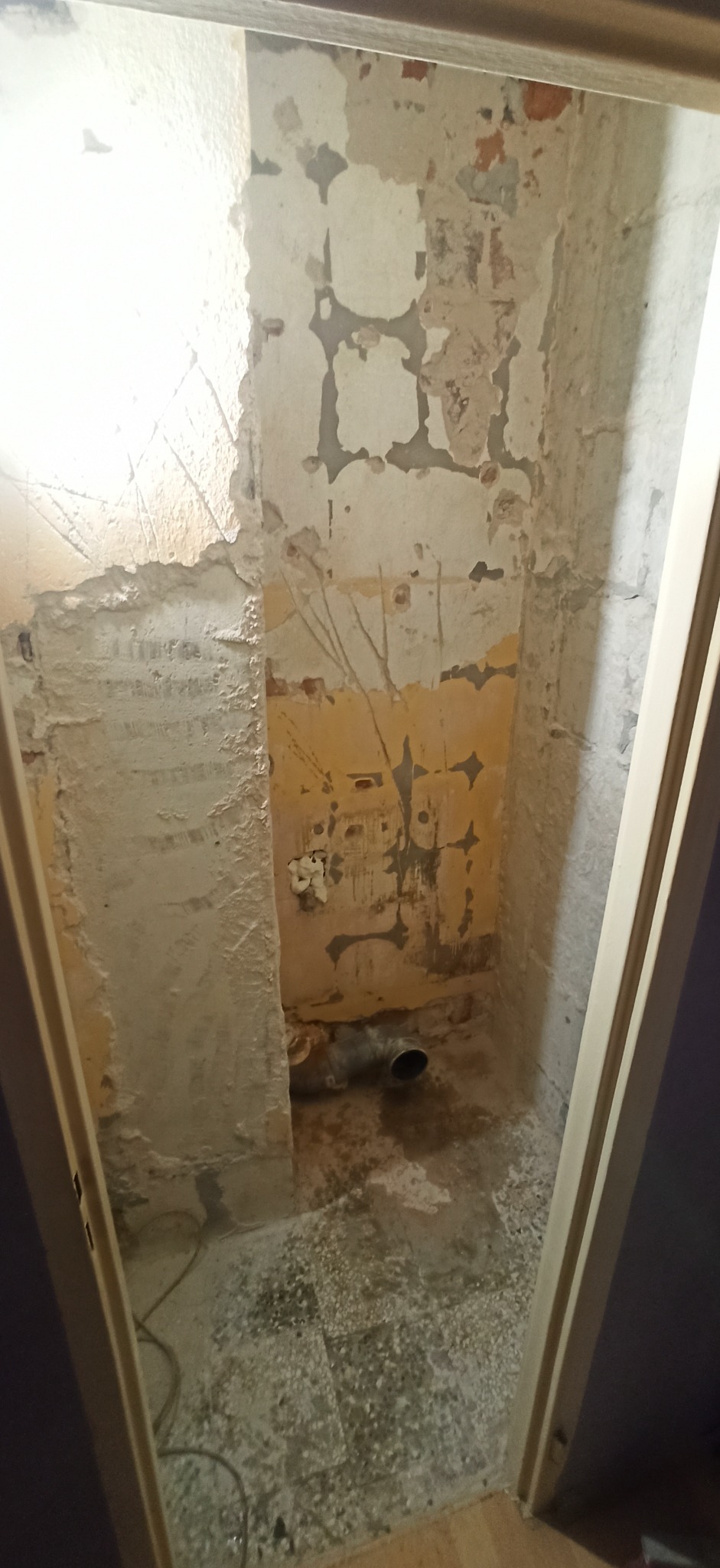 Wnętrze łazienki w trakcie demontażu, widoczne ściany z resztkami starej farby i tynku, odsłonięta instalacja kanalizacyjna, podłoga pokryta gruzem i pozostałościami po płytkach.