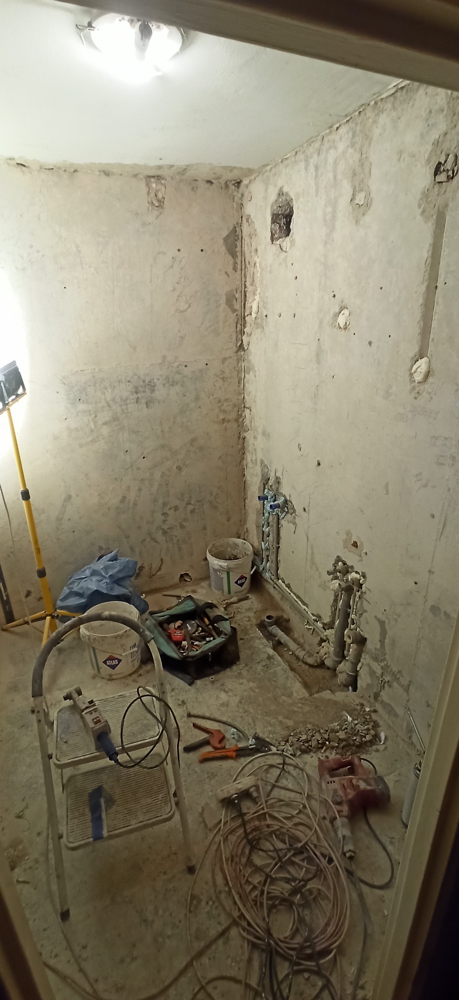 Łazienka w trakcie remontu: odsłonięte rury, gruz na podłodze, drabina i narzędzia, widoczne ślady po skuciu płytek.
