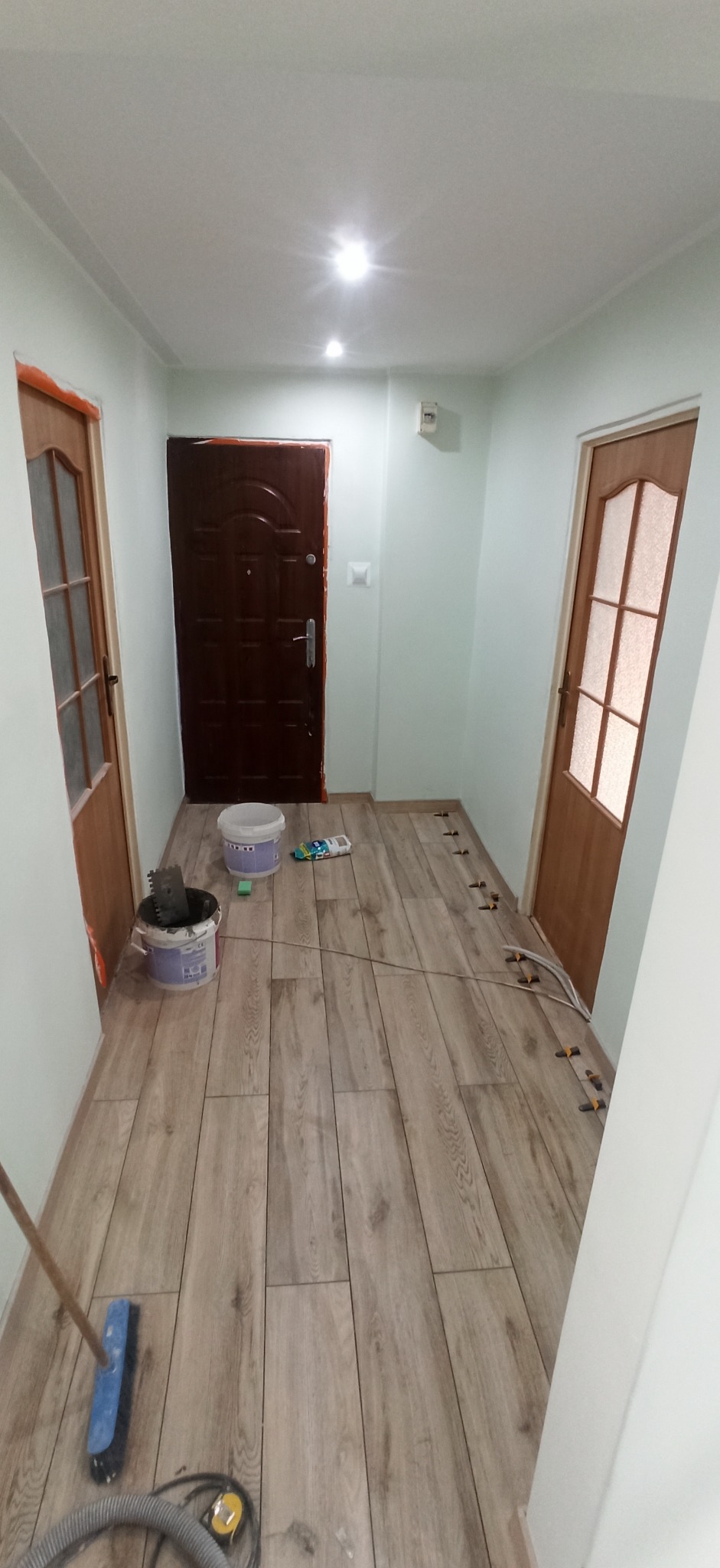 Korytarz w trakcie układania podłogi z płytek imitujących drewno w Warce. Widoczne wiadra z zaprawą, szpachelka, krzyżyki dystansowe i odkurzacz. Częściowo ułożona podłoga z fugami.