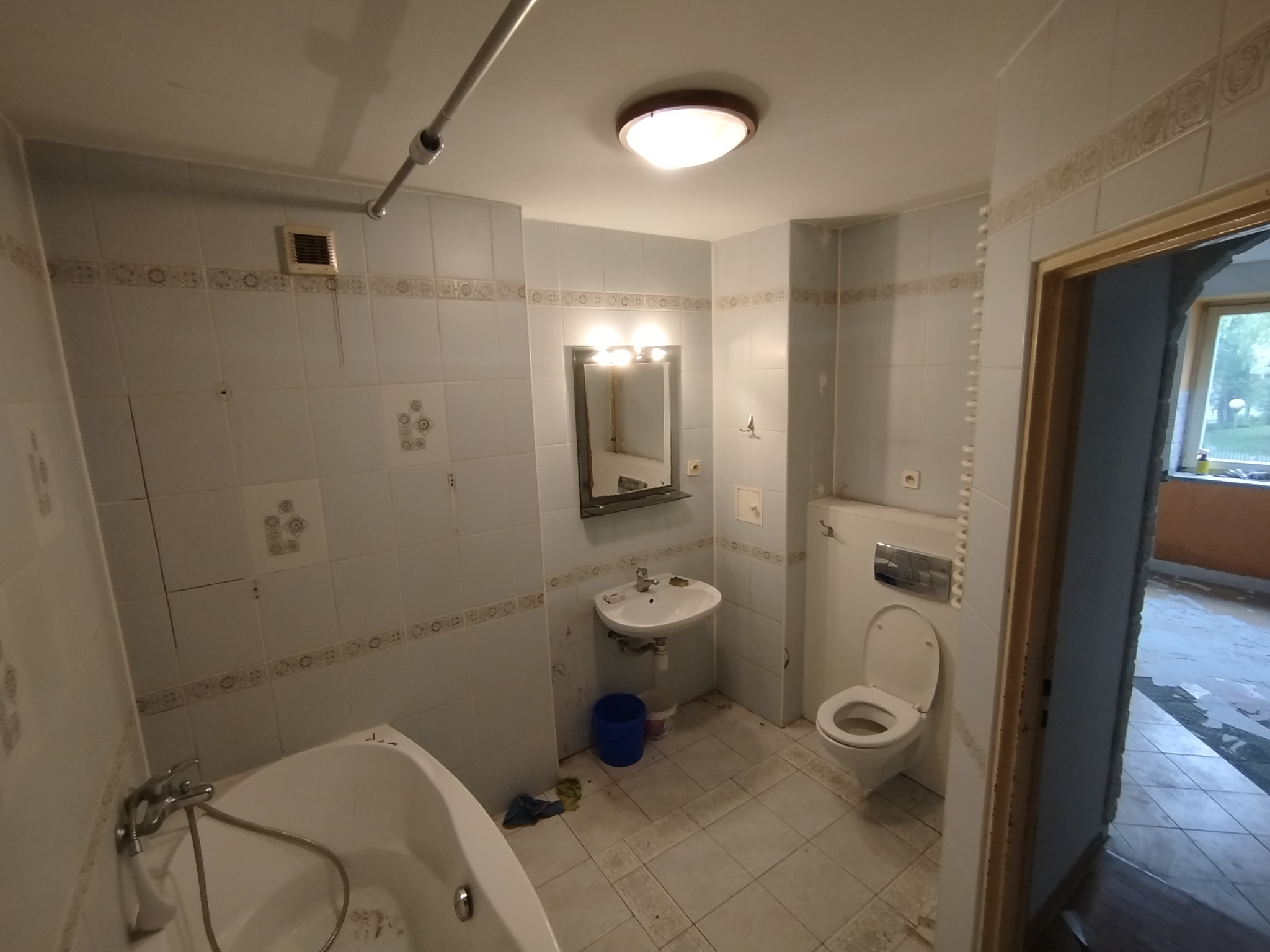 Łazienka w trakcie demontażu: widoczna wanna, umywalka z lustrem, toaleta, zdemontowane płytki na ścianach i podłodze, widoczne ślady po kleju i gruz.