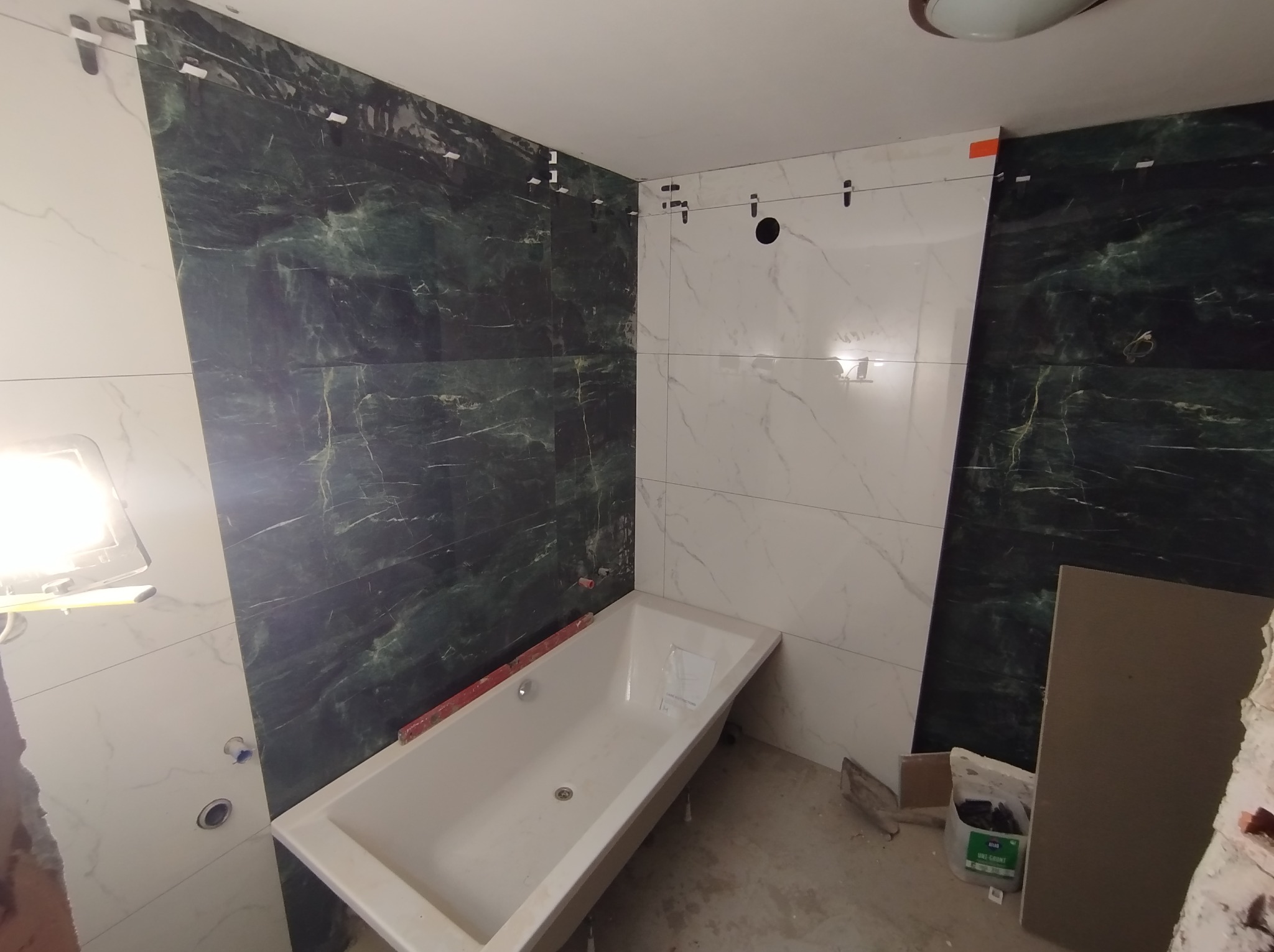 Remont łazienki: montaż płytek imitujących marmur w odcieniach bieli i zieleni, widoczna wanna i poziomica, etap prac wykończeniowych.