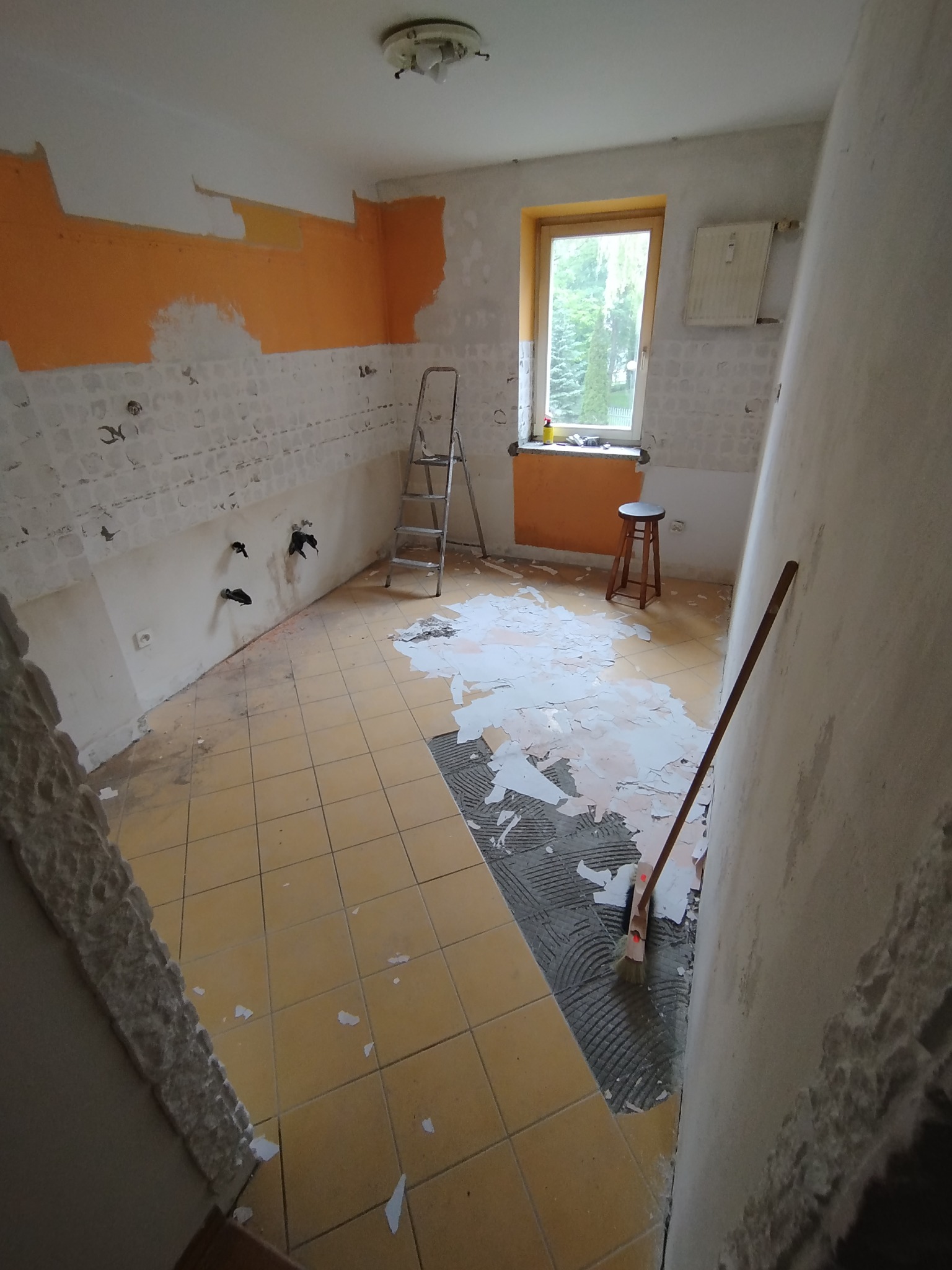 Pomieszczenie w trakcie demontażu płytek ściennych i podłogowych, widoczna drabina, resztki kleju i farby na ścianach, odsłonięte przyłącza hydrauliczne.