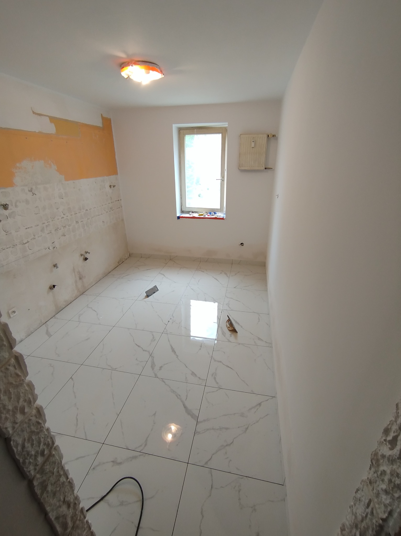 Wnętrze łazienki w trakcie remontu z ułożonymi białymi płytkami imitującymi marmur na podłodze, częściowo przygotowaną ścianą pod glazurę i widocznym grzejnikiem pod oknem.