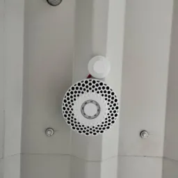 Biały czujnik dymu z wzorem w kształcie plastra miodu zamontowany na białym, panelowym suficie, widoczny również fragment szarej rury wentylacyjnej.