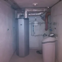 Szara pompa ciepła z widocznymi rurami instalacji w małym pomieszczeniu, obok zmiękczacz wody.