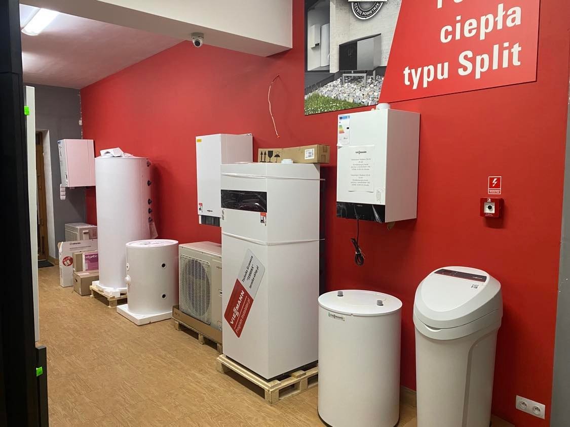 Ekspozycja różnorodnych urządzeń grzewczych, w tym zbiorników i jednostek zewnętrznych, ustawionych w rzędzie na drewnianych paletach wewnątrz pomieszczenia z czerwoną ścianą. Widoczna reklama...