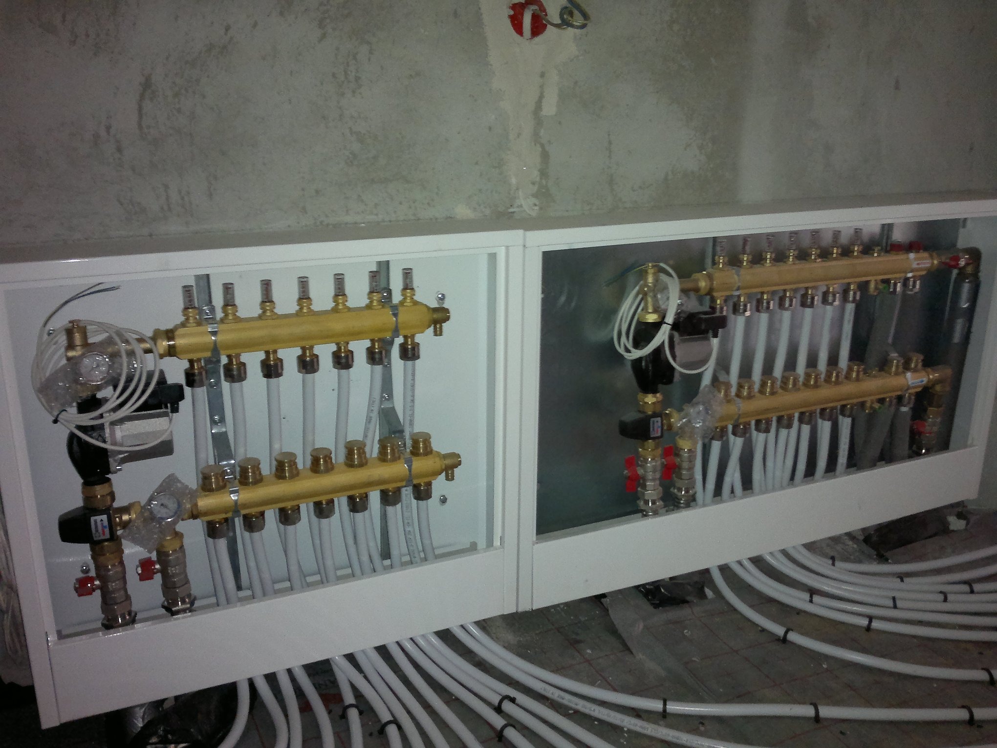 Rozdzielacz ogrzewania podłogowego w białej obudowie, z widocznymi mosiężnymi elementami, rurami i przewodami, zamontowany na tle surowej, szarej ściany.