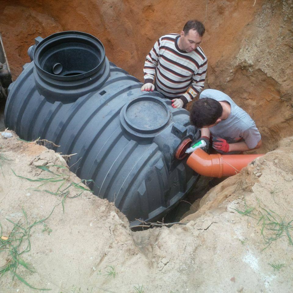 Montaż szarej, żebrowanej, plastikowej oczyszczalni ścieków w wykopie ziemnym; dwaj mężczyźni w roboczych ubraniach uszczelniają pomarańczową rurę odpływową.
