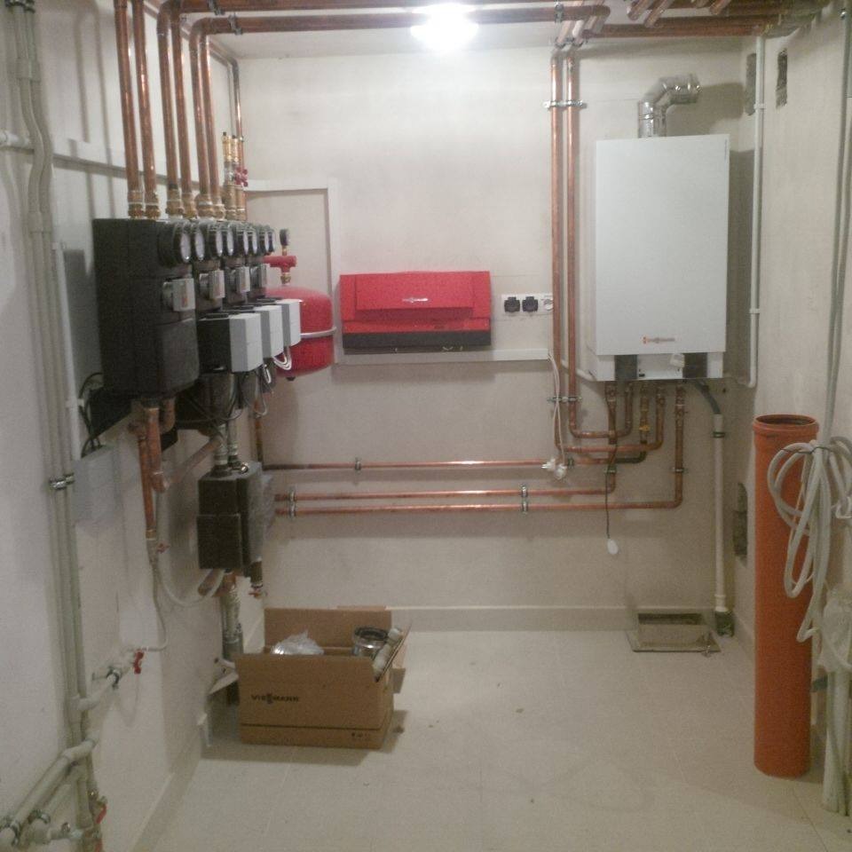 Kompletna instalacja centralnego ogrzewania w kotłowni: biały piec gazowy, czerwony zasobnik, czarne rozdzielacze, miedziane rury, karton Viessmann na podłodze.