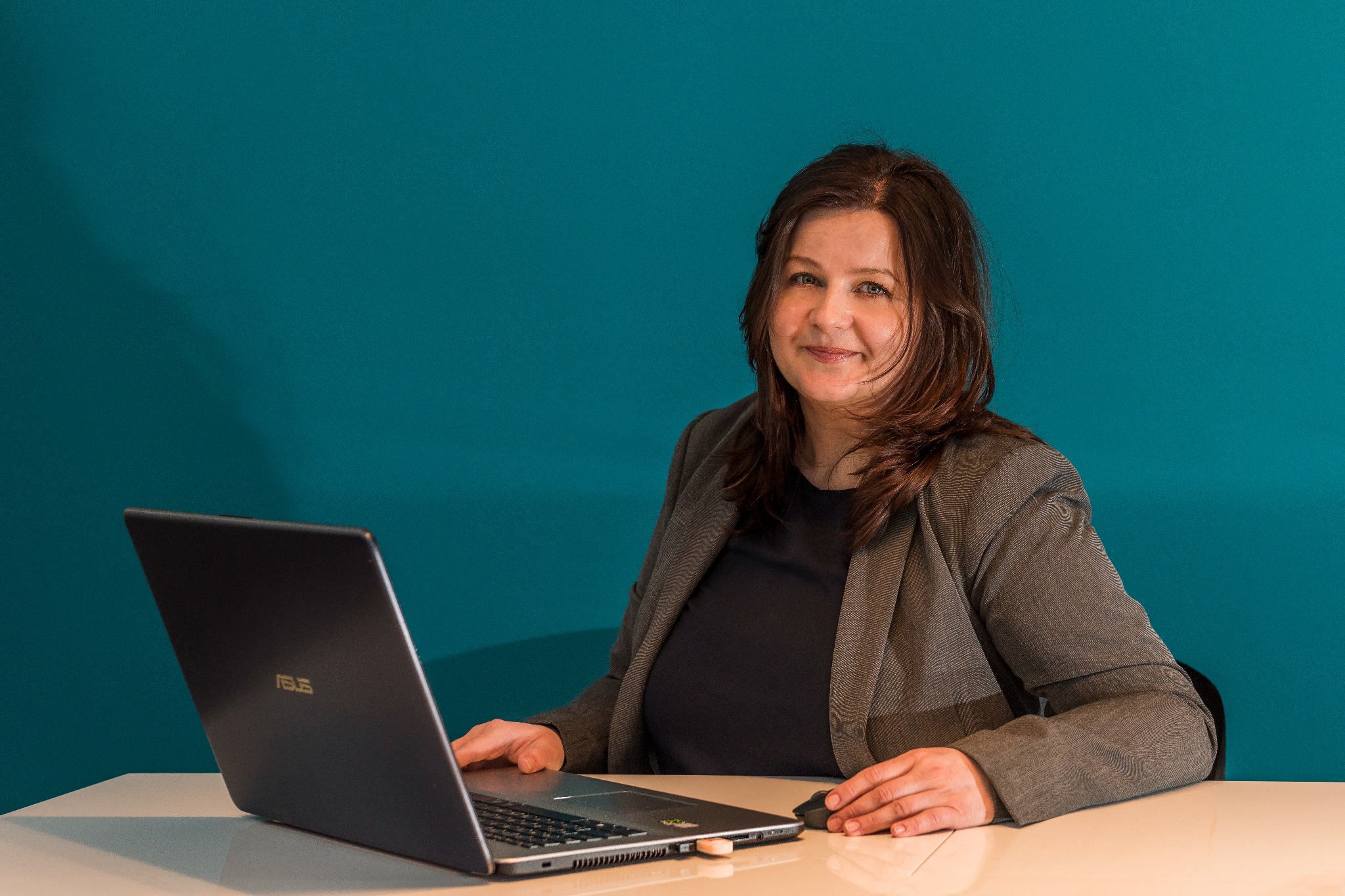 Uśmiechnięta kobieta w marynarce siedzi za biurkiem z laptopem Asus na tle turkusowej ściany.