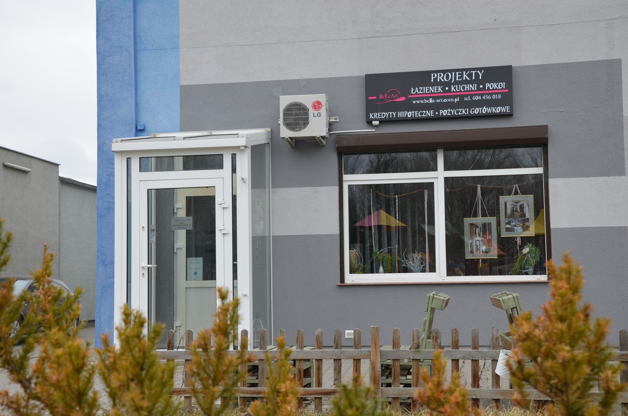 Fasada budynku usługowego z szyldem 'Projekty łazienek, kuchni, pokoi Bella Art', klimatyzatorem LG, oknem z dekoracjami i drewnianym płotem z roślinami na pierwszym planie.