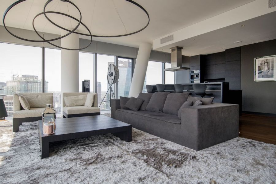Nowoczesny salon z szarym narożnikiem, dwoma fotelami, stolikiem kawowym i puszystym dywanem na tle panoramicznych okien z widokiem na miasto i otwartej kuchni z czarnymi szafkami.