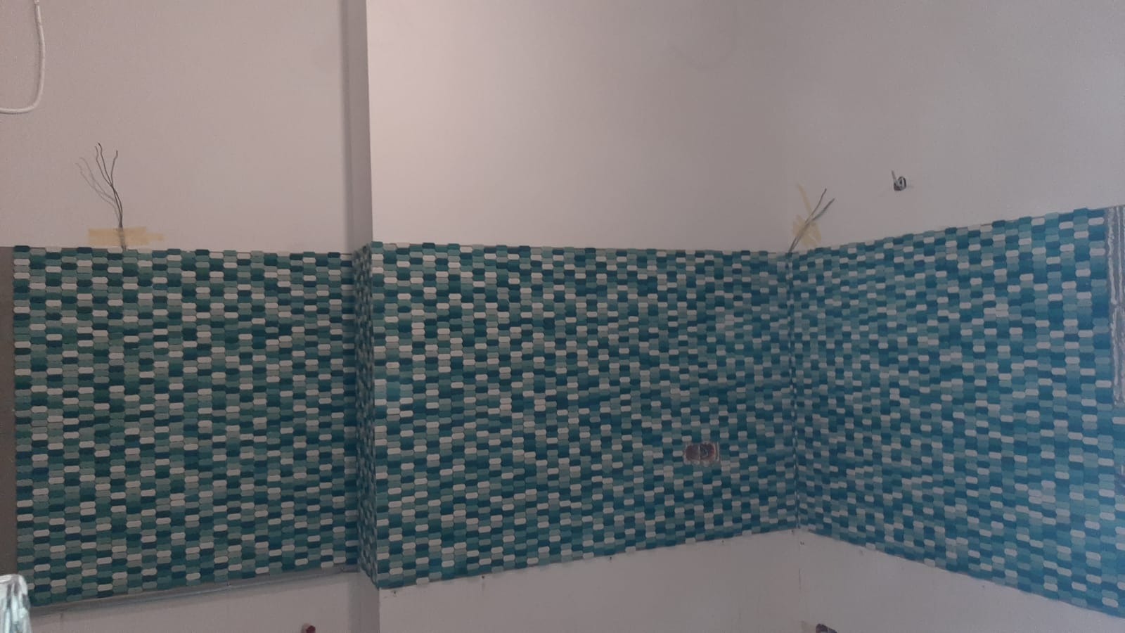 Ściana w trakcie układania mozaiki z drobnych, turkusowo-białych płytek w kształcie zaokrąglonych prostokątów, widoczne przewody elektryczne wystające ze ściany powyżej płytek.