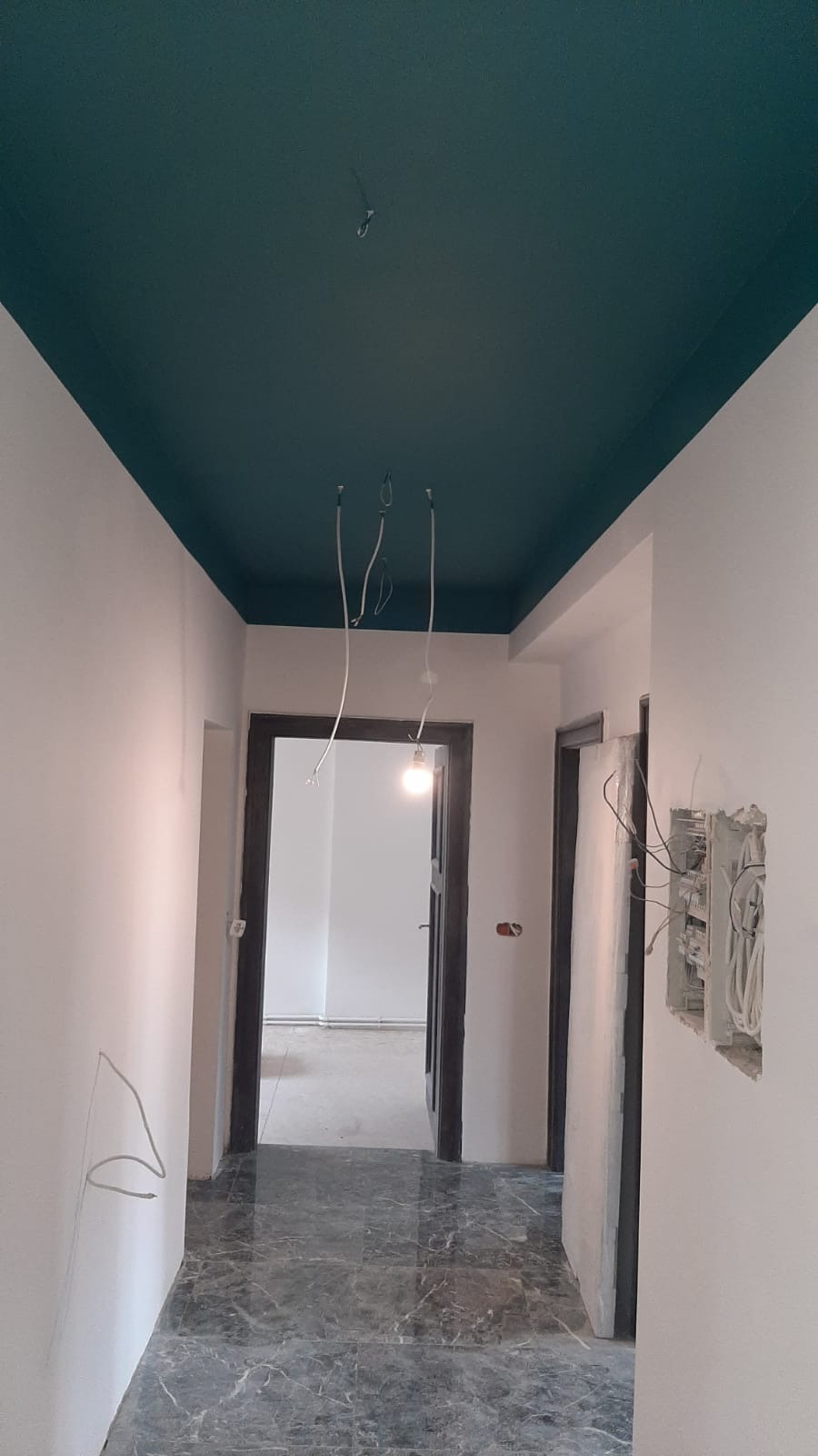 Korytarz w trakcie prac wykończeniowych: pomalowany na ciemnozielono sufit, białe ściany, marmurowe płytki na podłodze, wystające kable elektryczne, widoczna skrzynka elektryczna i drzwi do innych...