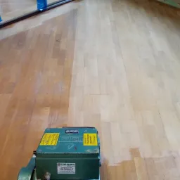 Parkiet w trakcie renowacji, widoczna r&oacute;żnica w kolorze między fragmentem po obr&oacute;bce a surowym drewnem, maszyna do szlifowania podł&oacute;g w dolnej części kadru.