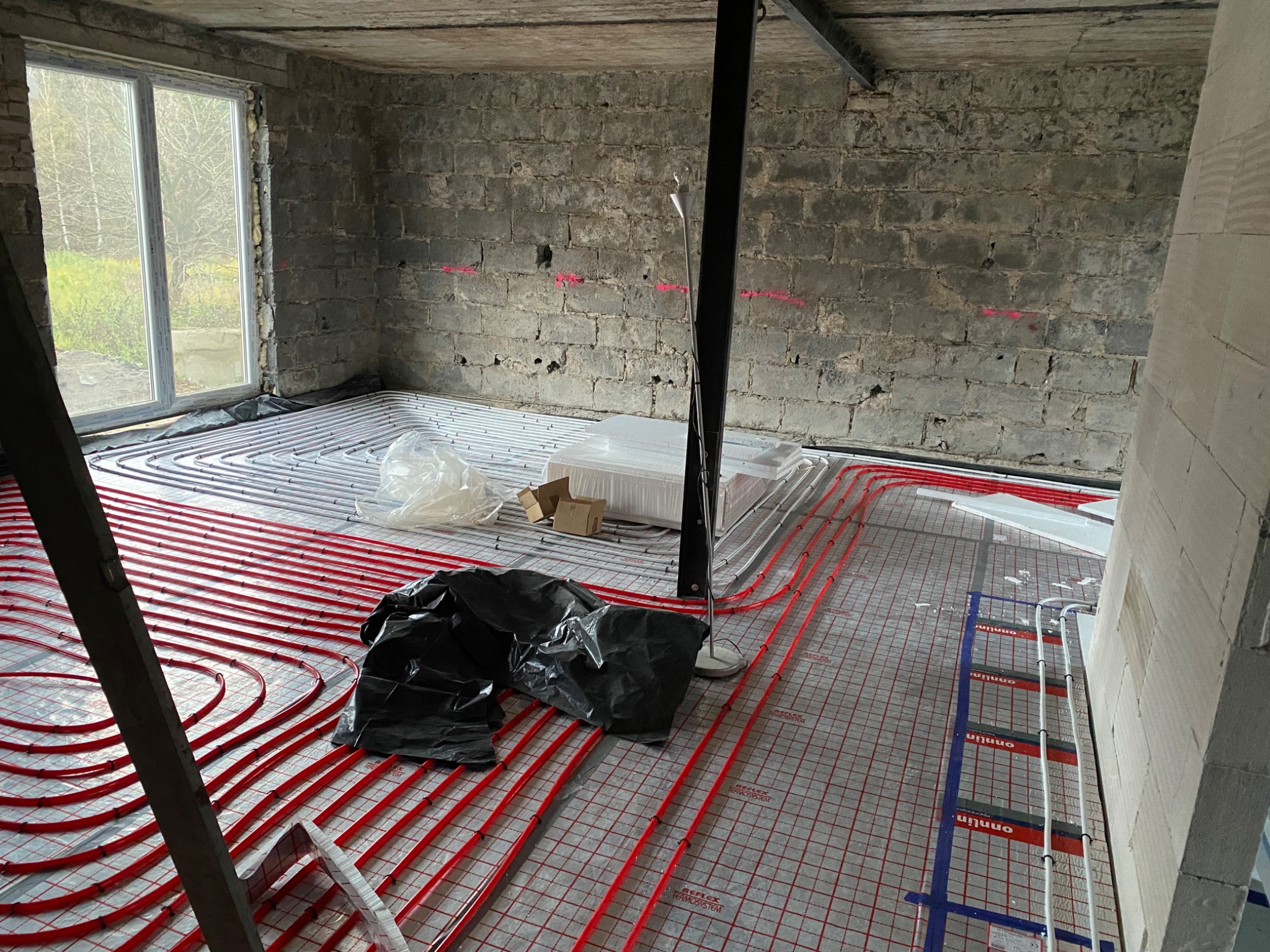 Rozłożone rury ogrzewania podłogowego w surowym pomieszczeniu z ceglanymi ścianami i dużym oknem, widoczne materiały budowlane i narzędzia.