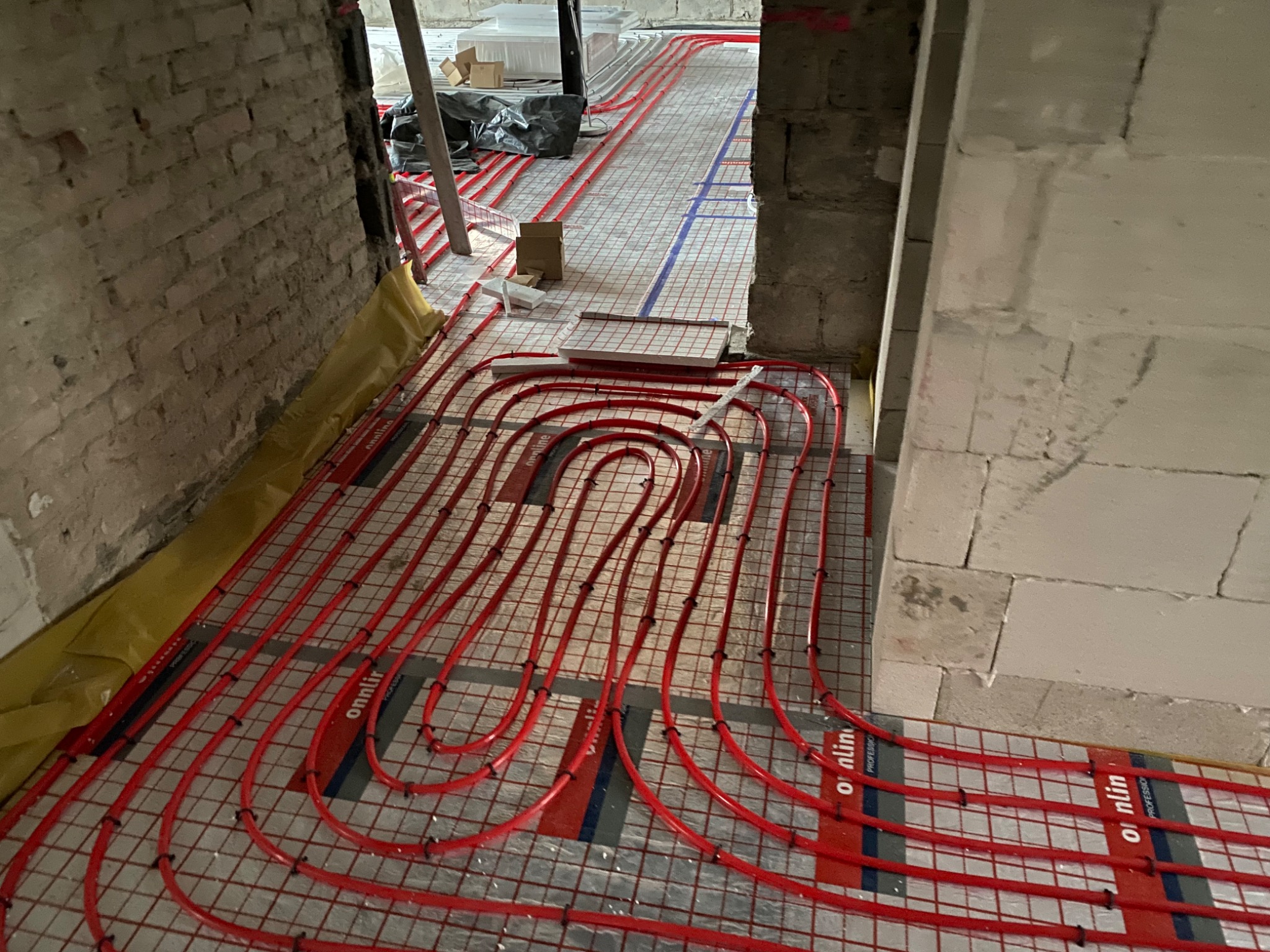 Montaż ogrzewania podłogowego z czerwonych rurek na siatce w surowym wnętrzu z ceglanymi ścianami i bloczkami betonowymi.