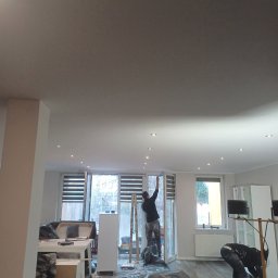 Andrzej  Rzepka - Prace malarskie w trakcie wykonywania w mieszkaniu w Głogowie: malarz na drabinie maluje sufit w pomieszczeniu z oknami, drugi malarz pochylony nad podłogą zabezpieczoną folią.