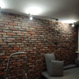 Andrzej  Rzepka - Ściana z cegły dekoracyjnej w salonie, oświetlona lampkami punktowymi, widoczna drabina malarska i fragment sofy z poduszką, na podłodze wiadro z farbą.