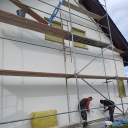 Andrzej  Rzepka - Dwóch pracowników na rusztowaniu wyrównuje świeżo nałożony tynk na elewacji domu w Głogowie, widoczne narzędzia i przygotowane okna.