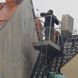 Pracownik na podnośniku koszowym czyści elewację budynku za pomocą myjki ciśnieniowej, w tle widoczny komin z czerwonej cegły i dach pokryty dachówką.