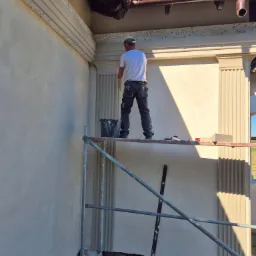 Pracownik na rusztowaniu, w trakcie nakładania zaprawy na elewację budynku z dekoracyjnymi elementami architektonicznymi, widoczny kubeł z zaprawą i poziomica.