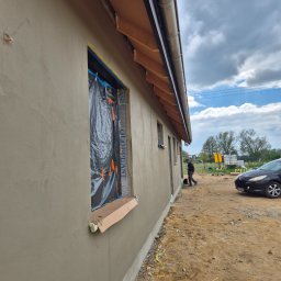 Ogolnobudowlana - Świeżo otynkowana elewacja budynku z zabezpieczonymi oknami folią i listwami. Widoczny fragment dachu, rynna, pracownik w oddali i zaparkowany samochód.