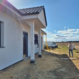 Ogolnobudowlana - Dom jednorodzinny w trakcie prac wykończeniowych elewacji. Widoczny pracownik na drabinie, biała elewacja, błękitne niebo i fragment krajobrazu.