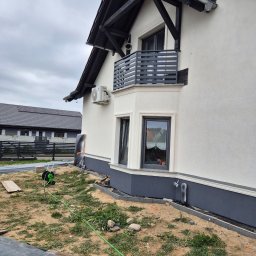 Ogolnobudowlana - Biała elewacja domu z balkonem i czarnymi elementami wykończeniowymi. Widoczny klimatyzator, trawa i narzędzia ogrodowe. Ujęcie z dołu, pochmurne niebo.