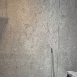 Ogolnobudowlana - Szara ściana z efektem betonu, widoczna fuga pionowa, fragment mopa i sterta ubrań w rogu. Minimalistyczne wykończenie wnętrza, surowy styl.