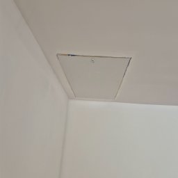 Ogolnobudowlana - Biały sufit z zamontowaną klapą rewizyjną, widok z dołu. Klapa z delikatnym obramowaniem, wtopiona w gładką powierzchnię sufitu. Minimalistyczny design.