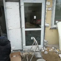 Montaż okien i ocieplenie elewacji budynku jednorodzinnego w trakcie realizacji. Widoczne białe okna PCV z pianką montażową, styropian na ścianach, drabina i narzędzia.