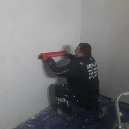 Pracownik w czarnej bluzie z logo firmy nakłada czerwoną pacą masę szpachlową na białą ścianę w rogu pokoju, widoczna drabina malarska i zabezpieczona folią podłoga.