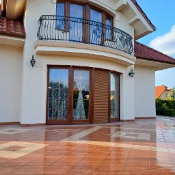 Jasna elewacja domu z balkonem z kutą balustradą, widoczne drewniane okna z firankami i płytki na tarasie z odbiciami światła.