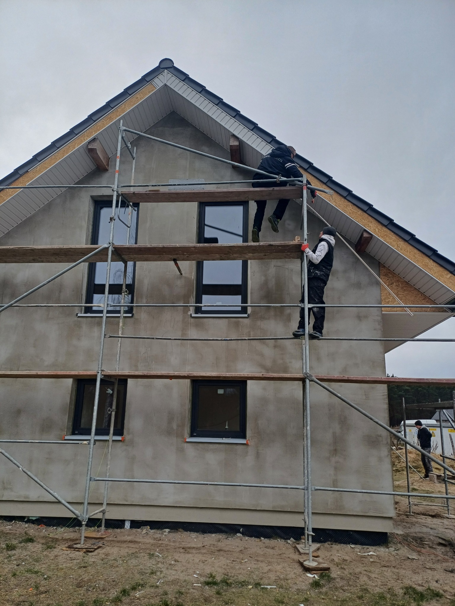 Dwaj robotnicy na rusztowaniu pracują przy elewacji domu jednorodzinnego z szarą, surową powierzchnią, widoczne czarne okna i drewniane elementy konstrukcyjne dachu.