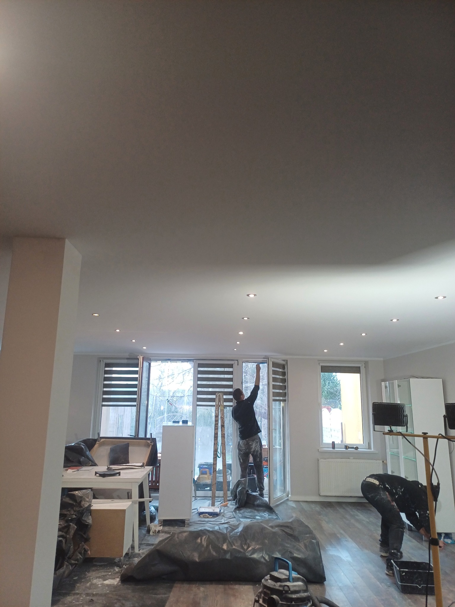 Prace malarskie w trakcie wykonywania w mieszkaniu w Głogowie: malarz na drabinie maluje sufit w pomieszczeniu z oknami, drugi malarz pochylony nad podłogą zabezpieczoną folią.